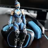 A detailed action figure dressed in a futuristic blue and black outfit sits atop a gaming headset. The figure features armor-like components with intricate designs and a mask covering part of its face. The headset is predominantly black with blue padding and accents, positioned near a computer monitor.