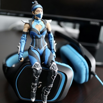 A detailed action figure dressed in a futuristic blue and black outfit sits atop a gaming headset. The figure features armor-like components with intricate designs and a mask covering part of its face. The headset is predominantly black with blue padding and accents, positioned near a computer monitor.