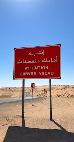 A red road sign with Arabic and English text warning of curves ahead is prominently displayed. The sign is situated in a desert landscape with a paved road curving through sandy dunes under a clear blue sky.