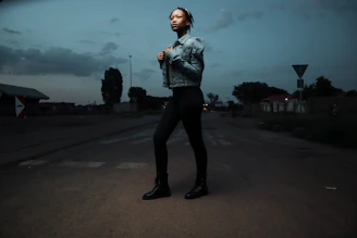 A stylish man wearing a sleek jacket and jeans, standing confidently on a city street at dusk.
