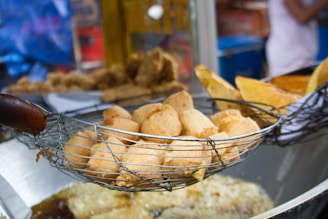 A close-up view of a batch of golden-brown fried snacks resting in a metal wire skimmer. The background reveals a busy street food stall setting with slightly blurred elements, showcasing different types of fried foods.