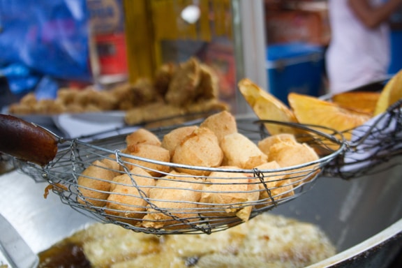 A close-up view of a batch of golden-brown fried snacks resting in a metal wire skimmer. The background reveals a busy street food stall setting with slightly blurred elements, showcasing different types of fried foods.