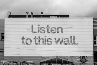 A large wall with horizontal paneling features the bold text 'Listen to this wall.' The wall is part of a building with visible rooftops and pipes. Graffiti is present at the bottom of the wall.