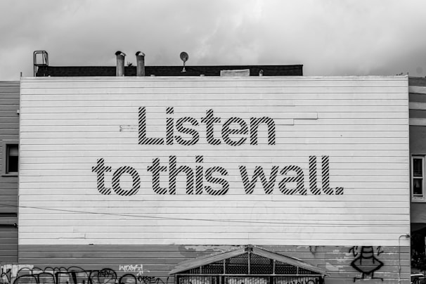 A large wall with horizontal paneling features the bold text 'Listen to this wall.' The wall is part of a building with visible rooftops and pipes. Graffiti is present at the bottom of the wall.