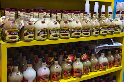 Shelves filled with multiple bottles of cream liqueurs, labeled 'Fruitcream,' arranged neatly on a bright yellow display in a store. Various flavors are indicated such as piña and banana. Disposable cups are stacked on the top shelf, and newspapers are stacked on the lower shelf.