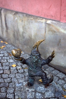 A small gnome-like statue made of bronze stands on a cobblestone walkway. The figure has a pointy hat and beard, and it is holding a golden-colored object shaped like a heart with an emblem. The background includes a stone wall with a pinkish hue.