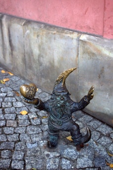 A small gnome-like statue made of bronze stands on a cobblestone walkway. The figure has a pointy hat and beard, and it is holding a golden-colored object shaped like a heart with an emblem. The background includes a stone wall with a pinkish hue.