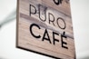 A wooden sign displaying the text 'PURO CAF&Eacute;' with a rustic, natural appearance. The background is a light wood texture with visible grains, and the font is bold in black.