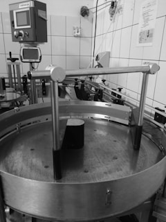 An industrial setting with machinery including a circular metal platform and electronic control panels mounted on a tiled wall. Various pipes and wires run along the walls and ceiling, with different mechanical components visible.