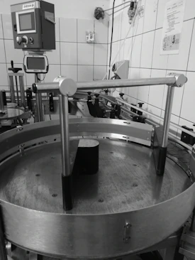 An industrial setting with machinery including a circular metal platform and electronic control panels mounted on a tiled wall. Various pipes and wires run along the walls and ceiling, with different mechanical components visible.