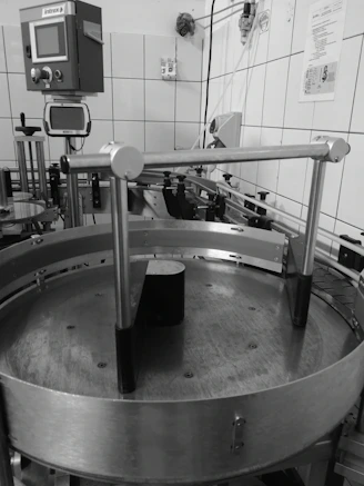 An industrial setting with machinery including a circular metal platform and electronic control panels mounted on a tiled wall. Various pipes and wires run along the walls and ceiling, with different mechanical components visible.