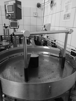 An industrial setting with machinery including a circular metal platform and electronic control panels mounted on a tiled wall. Various pipes and wires run along the walls and ceiling, with different mechanical components visible.