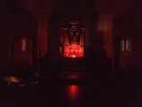 A dimly lit interior space with a central altar-like structure illuminated by a red light. The room appears to be a chapel or religious setting with dark wooden furniture and stained-glass windows on either side.