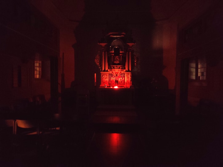 A dimly lit interior space with a central altar-like structure illuminated by a red light. The room appears to be a chapel or religious setting with dark wooden furniture and stained-glass windows on either side.