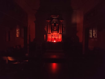 A dimly lit interior space with a central altar-like structure illuminated by a red light. The room appears to be a chapel or religious setting with dark wooden furniture and stained-glass windows on either side.
