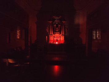 A dimly lit interior space with a central altar-like structure illuminated by a red light. The room appears to be a chapel or religious setting with dark wooden furniture and stained-glass windows on either side.