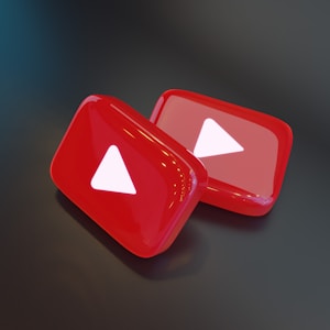 Two shiny, red, three-dimensional shapes resembling play buttons are positioned on a dark surface. Each button displays a white triangle in the center, which is characteristic of a popular video sharing platform.