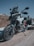 A modern adventure motorcycle is parked on a gravel path, showcasing a dynamic and rugged design. The bike features a large windshield, robust frame, and dual-sport tires, indicating its capability for both on-road and off-road riding. The setting includes a clear blue sky and a rocky terrain in the background.
