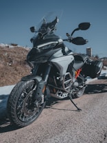 A modern adventure motorcycle is parked on a gravel path, showcasing a dynamic and rugged design. The bike features a large windshield, robust frame, and dual-sport tires, indicating its capability for both on-road and off-road riding. The setting includes a clear blue sky and a rocky terrain in the background.