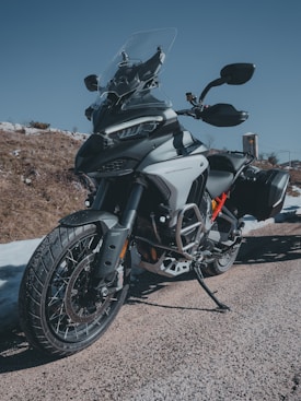 A modern adventure motorcycle is parked on a gravel path, showcasing a dynamic and rugged design. The bike features a large windshield, robust frame, and dual-sport tires, indicating its capability for both on-road and off-road riding. The setting includes a clear blue sky and a rocky terrain in the background.