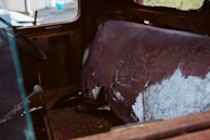 The image depicts the interior of a deteriorated vehicle with a torn, faded seat and rusted metal surfaces. The seat's upholstery is severely damaged, exposing the underlying material. The steering wheel is intact, though showing signs of age, and the gear shift lever is visible on the left side.
