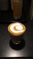 A small glass of coffee topped with a swirl of milk foam is placed on a dark countertop. In the background, there is a bottle labeled 'Caramel Drizzle'.