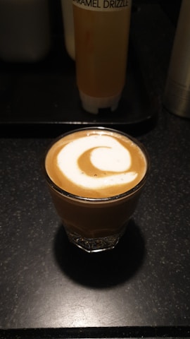 A small glass of coffee topped with a swirl of milk foam is placed on a dark countertop. In the background, there is a bottle labeled 'Caramel Drizzle'.