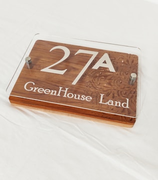 A rectangular plaque made of wood with the text '27A GreenHouse Land' inscribed. It has a transparent acrylic cover secured with two metallic bolts at the corners. The background is a plain white surface.