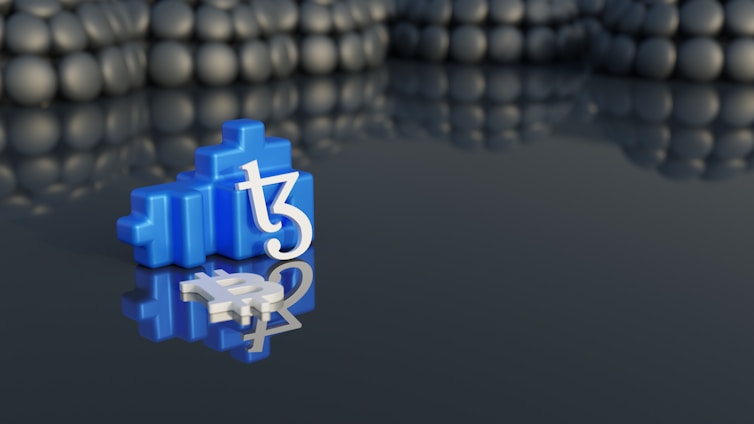 A blue, block-like structure featuring the Tezos (XTZ) logo stands prominently on a reflective surface, creating a mirror image. The background is composed of blurry, rounded shapes that resemble stones or pearls.
