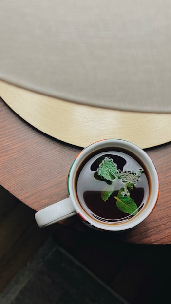 A cozy wooden table with a steaming cup of herbal tea surrounded by fresh green leaves and dried tea herbs.