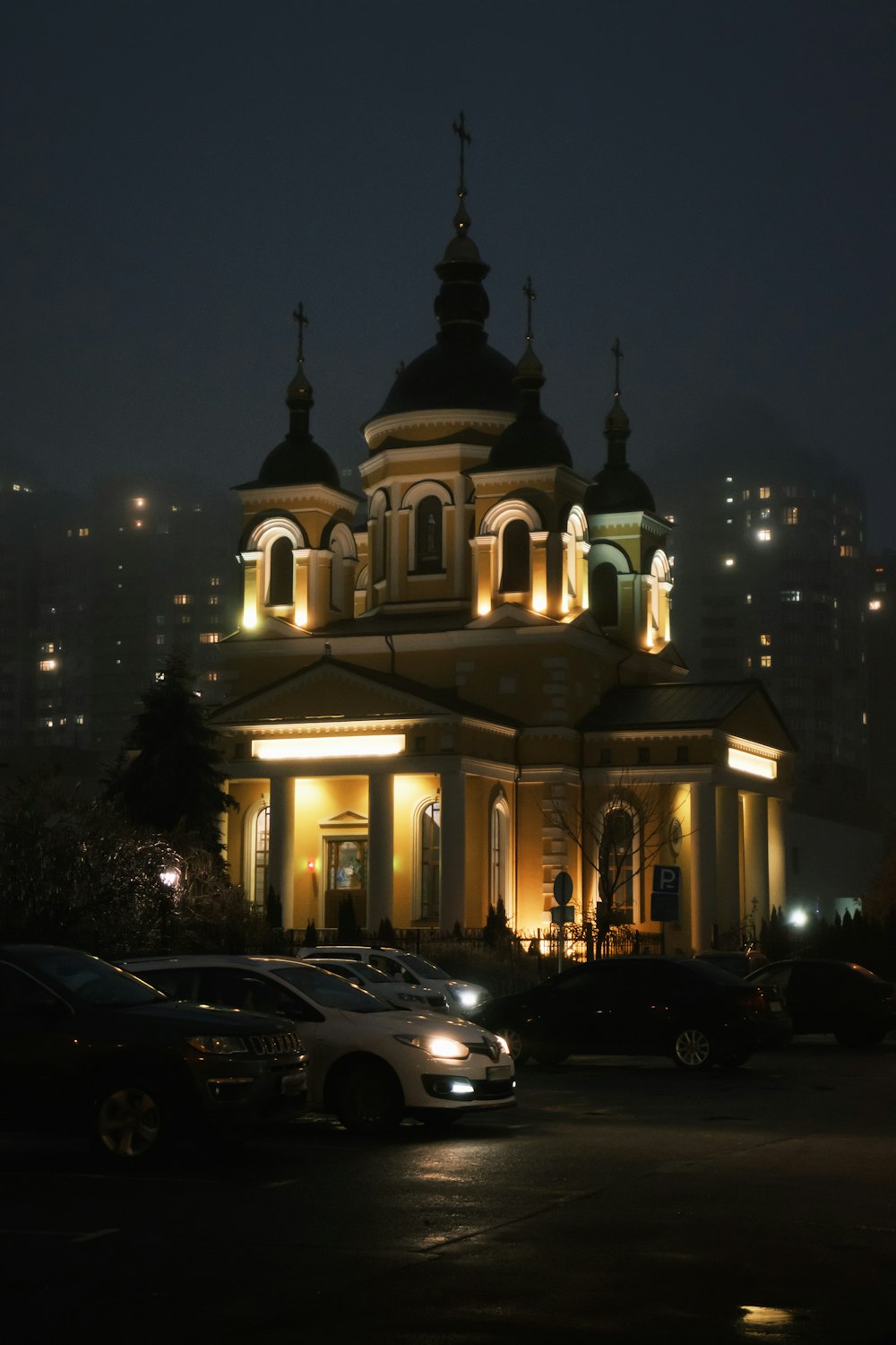 uma igreja iluminada à noite com carros estacionados em frente a ela