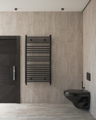 A modern bathroom showcases a minimalist design with light wooden panel walls and flooring. A sleek black wall-mounted toilet is on the right, complemented by a black heated towel rail in the center. The wall on the left features a door with horizontal slats for a contemporary aesthetic.