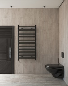 A modern bathroom showcases a minimalist design with light wooden panel walls and flooring. A sleek black wall-mounted toilet is on the right, complemented by a black heated towel rail in the center. The wall on the left features a door with horizontal slats for a contemporary aesthetic.
