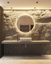 A contemporary bathroom features a round mirror with a backlit, textured wall resembling natural rock. Beneath the mirror, there is a sleek countertop with a modern, rectangular white sink and a minimalist black faucet. A small shelf holds several small bottles and reed diffusers, adding an element of sophistication.