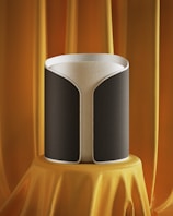 A modern, cylindrical speaker with a sleek design is displayed on a pedestal draped in luxurious orange fabric. The speaker has a gray and black fabric exterior with a subtle texture and a white top edge.