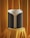 A modern, cylindrical speaker with a sleek design is displayed on a pedestal draped in luxurious orange fabric. The speaker has a gray and black fabric exterior with a subtle texture and a white top edge.