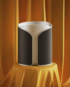 A modern, cylindrical speaker with a sleek design is displayed on a pedestal draped in luxurious orange fabric. The speaker has a gray and black fabric exterior with a subtle texture and a white top edge.