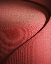 A detailed close-up of a red velvet or corduroy fabric arranged in an abstract, flowing pattern. The texture of the fabric is prominently visible, with fine horizontal lines creating a ribbed effect. The curvature suggests an artistic or furniture design component, possibly part of a sofa or chair.