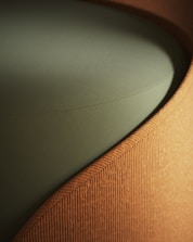 A close-up view of a piece of furniture with a textured, ribbed fabric surface in a warm, earthy brown color, forming a smooth, gently curving shape. The seat cushion is upholstered in a solid, muted green fabric, providing a contrasting texture and color.