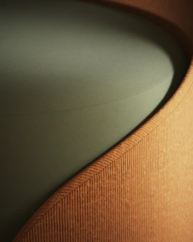 A close-up view of a piece of furniture with a textured, ribbed fabric surface in a warm, earthy brown color, forming a smooth, gently curving shape. The seat cushion is upholstered in a solid, muted green fabric, providing a contrasting texture and color.