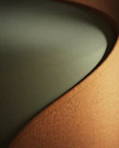 A close-up view of a piece of furniture with a textured, ribbed fabric surface in a warm, earthy brown color, forming a smooth, gently curving shape. The seat cushion is upholstered in a solid, muted green fabric, providing a contrasting texture and color.