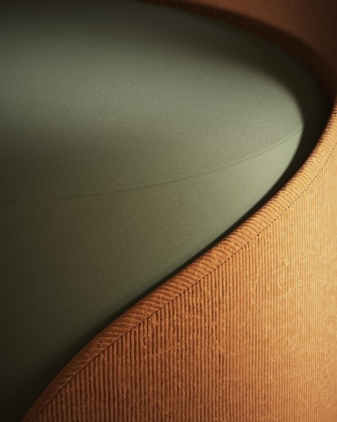 A close-up view of a piece of furniture with a textured, ribbed fabric surface in a warm, earthy brown color, forming a smooth, gently curving shape. The seat cushion is upholstered in a solid, muted green fabric, providing a contrasting texture and color.