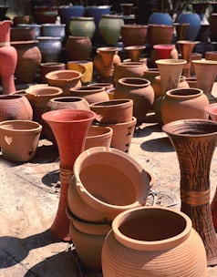 A variety of clay pots and vases are arranged outdoors on a sunlit surface. The pottery includes different shapes and sizes, ranging from wide, round pots to tall, slender vases. Some pots have intricate carvings and designs.