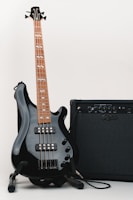 A black electric bass guitar with a brown fretboard stands on a stand next to a black amplifier. The guitar features four strings and multiple pickups, and the amplifier has a panel with various knobs and inputs.
