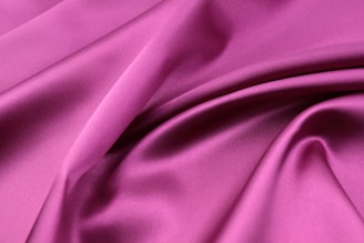 A smooth, lustrous piece of fabric in a rich pink hue. The fabric is draped in soft folds, showcasing its silky texture and gentle sheen.