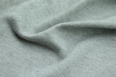 A close-up view of soft, ribbed fabric with a light green hue. The texture is detailed, revealing the individual knitting patterns with slight shadows and highlights, suggesting gentle folds and a smooth surface.