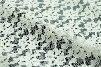 Intricate floral lace fabric with a delicate, sheer texture. The lace features an intricate pattern of flowers and leaves, with a mix of open and dense areas creating a sense of transparency.