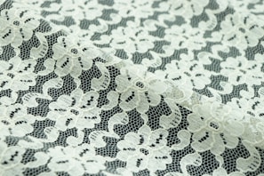 Intricate floral lace fabric with a delicate, sheer texture. The lace features an intricate pattern of flowers and leaves, with a mix of open and dense areas creating a sense of transparency.