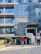 A modern building with a glass facade labeled as an Electronics Block. Several people are walking near the entrance. A person wearing a red sweater and jeans stands in the foreground on a tiled pathway. Landscaping accents including bushes are visible along the pathway.