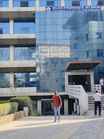 A modern building with a glass facade labeled as an Electronics Block. Several people are walking near the entrance. A person wearing a red sweater and jeans stands in the foreground on a tiled pathway. Landscaping accents including bushes are visible along the pathway.